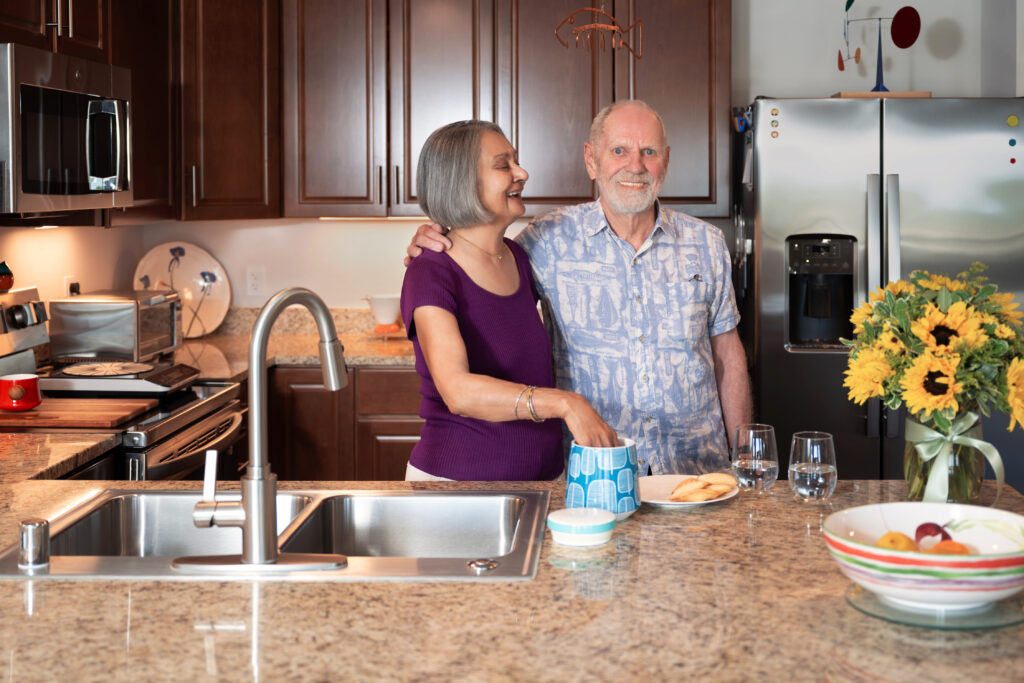 An older couple smiling in a modern kitchen as the woman reaches into a blue-patterned cookie jar on the marble countertop.